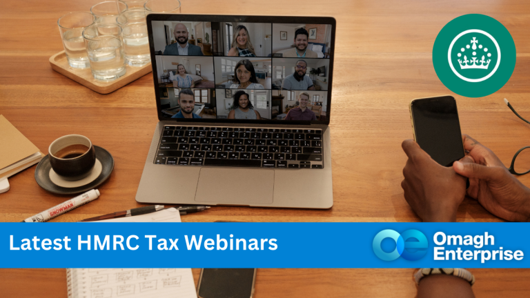 A laptop sitting on desk, with a webinar shown on screen. A hand shown, with mobile phone in it. A cup of tea, glasses of water, a notepad and pen, is on desk with reading glasses. A blue banner along the bottom, with white writing. "Latest HMRC Tax Webinars" Omagh Enterprise is on logo.