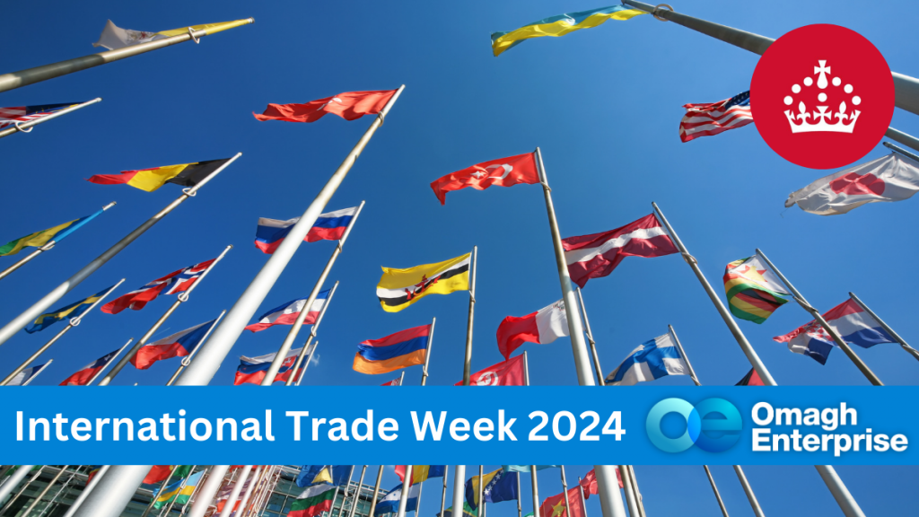 A group of international flags waving against a clear blue sky. A red circle with a white crown symbol is in the top right. A blue banner at the bottom reads International Trade Week 2024 with the Omagh Enterprise logo.